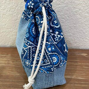 K&ouml;nnte beinhalten: Ein Kordelzugbeutel mit Jeansboden und blau gemustertem Paisley-Oberteil. Die Tasche ist mit einem wei&szlig;en Seil gebunden. Die Tasche ist aus Stoff und f&uuml;r kleine Gegenst&auml;nde konzipiert. Die Tasche steht auf einer Holzoberfl&auml;che.