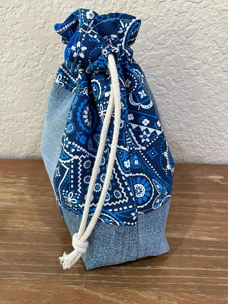 K&ouml;nnte beinhalten: Ein Kordelzugbeutel mit Jeansboden und blau gemustertem Paisley-Oberteil. Die Tasche ist mit einem wei&szlig;en Seil gebunden. Die Tasche ist aus Stoff und f&uuml;r kleine Gegenst&auml;nde konzipiert. Die Tasche steht auf einer Holzoberfl&auml;che.