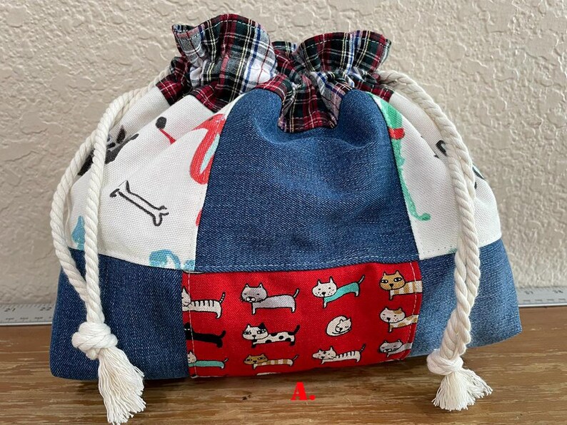 May include: A drawstring bag with a patchwork design. The bag features denim, white fabric with bone and red designs, and a red panel with cat illustrations. The top is plaid, and the drawstring is white.