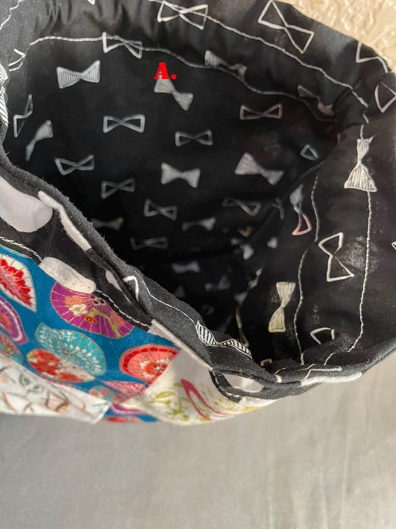 Pu&ograve; includere: Interno di una borsa in tessuto con sfondo nero e motivo a fiocco bianco. Il bordo della borsa presenta una stampa floreale e geometrica colorata. La fodera interna rivela il design a fiocco.