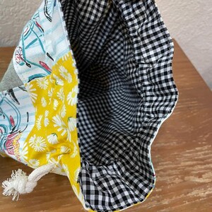 Op de afbeelding: Trekkoordtas met een patchwork ontwerp. De buitenkant heeft panelen met gele madeliefjesprint, fietsenprint en grijze stof. De binnenkant is gevoerd met zwart-wit geruite stof. Een wit touwtrekkoord is zichtbaar.