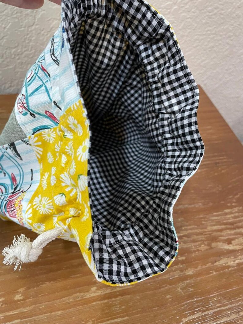 Op de afbeelding: Trekkoordtas met een patchwork ontwerp. De buitenkant heeft panelen met gele madeliefjesprint, fietsenprint en grijze stof. De binnenkant is gevoerd met zwart-wit geruite stof. Een wit touwtrekkoord is zichtbaar.