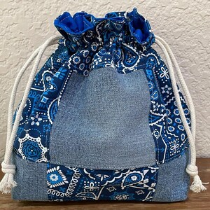 K&ouml;nnte beinhalten: Ein Kordelzugbeutel mit Jeans- und Paisley-Design. Die Tasche hat eine hellblaue Jeans-Mittelbahn, die von einem blau-wei&szlig;en Paisley-Muster umrahmt wird. Die Oberseite ist mit blauem Stoff und wei&szlig;em Kordelzug gerafft.