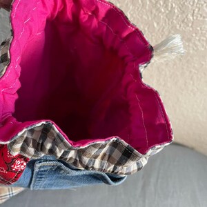 May include: Handmade drawstring bag with a bright pink interior. The exterior features a patchwork design with denim, plaid, and red patterned fabric. A white tassel is attached to the drawstring closure. The bag is open, revealing the interior.