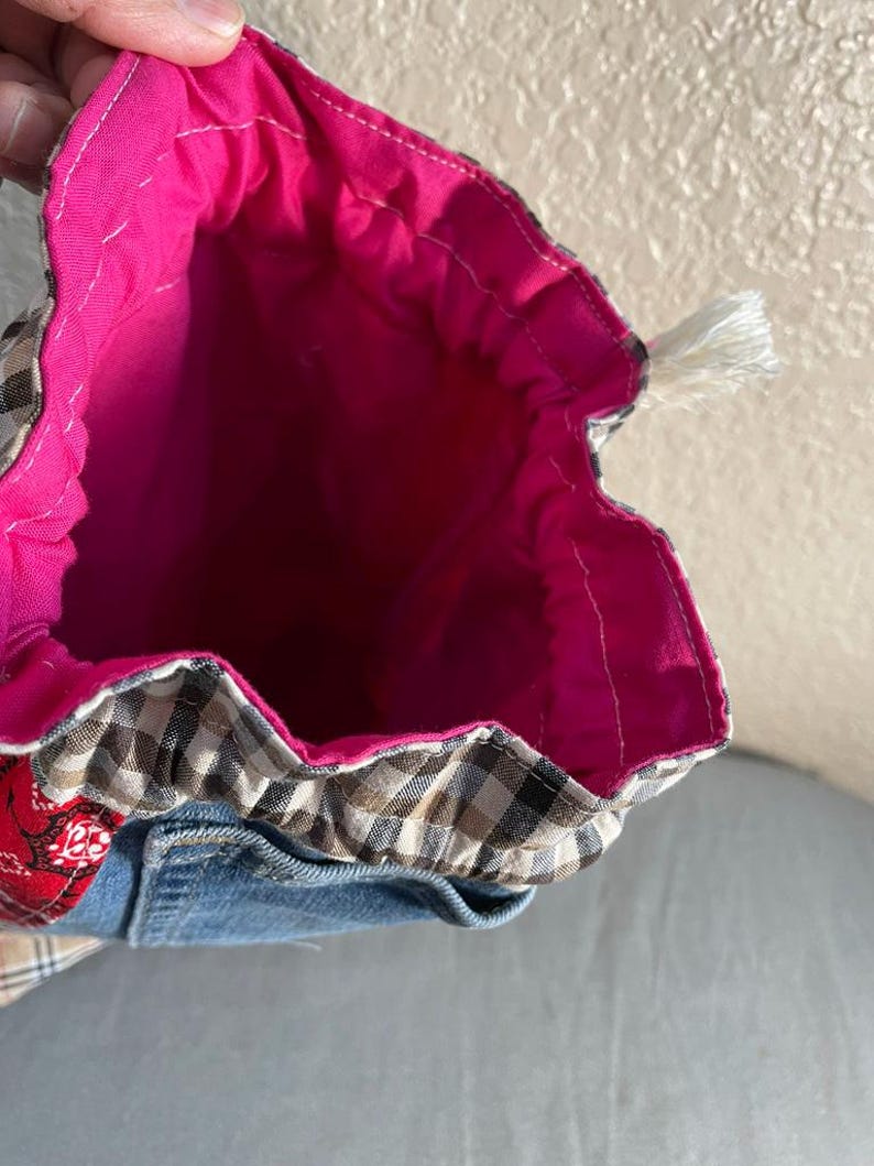 May include: Handmade drawstring bag with a bright pink interior. The exterior features a patchwork design with denim, plaid, and red patterned fabric. A white tassel is attached to the drawstring closure. The bag is open, revealing the interior.