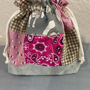 May include: A patchwork drawstring bag with a grey base and various patterned fabrics. The bag features pink, brown, and grey sections with bee, paisley, and dog bone designs. A white drawstring cord cinches the top.