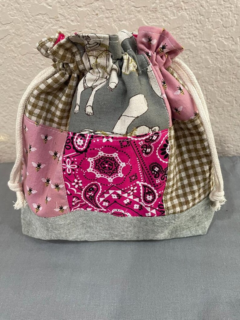 May include: A patchwork drawstring bag with a grey base and various patterned fabrics. The bag features pink, brown, and grey sections with bee, paisley, and dog bone designs. A white drawstring cord cinches the top.
