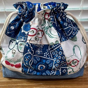 May include: A drawstring bag with a patchwork design. The bag features a denim base and a body composed of various fabric patches in white and blue, adorned with dog illustrations and paisley patterns. A cream-colored drawstring is visible.
