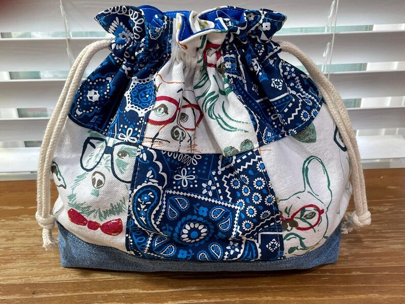 May include: A drawstring bag with a patchwork design. The bag features a denim base and a body composed of various fabric patches in white and blue, adorned with dog illustrations and paisley patterns. A cream-colored drawstring is visible.