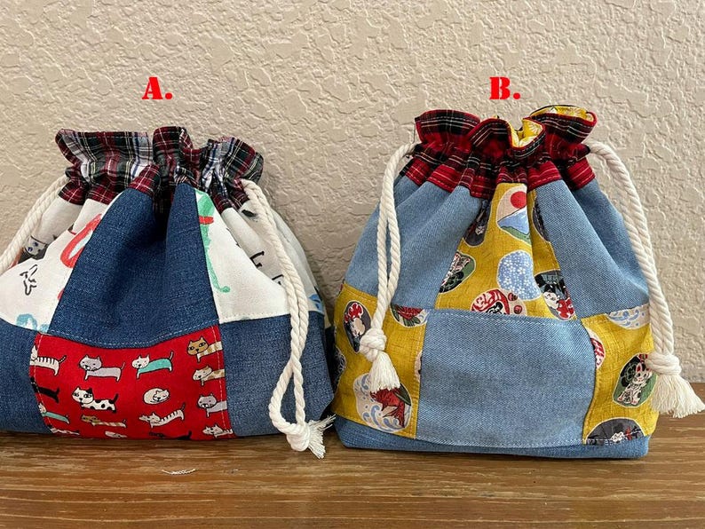 May include: Two drawstring bags with patchwork designs. One bag features a red panel with cat illustrations, while the other has a yellow panel with Japanese-inspired motifs. Both bags have denim and patterned fabric panels, with a plaid fabric at the top.