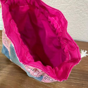 K&ouml;nnte beinhalten: Offener Kordelzugbeutel mit pinkfarbenem Innenraum und gemustertem Au&szlig;enbereich. Die Au&szlig;enseite weist ein paisleyartiges Design in Pink und Blau auf. Der Innenraum der Tasche ist leuchtend pink, und der Kordelzug ist wei&szlig;.