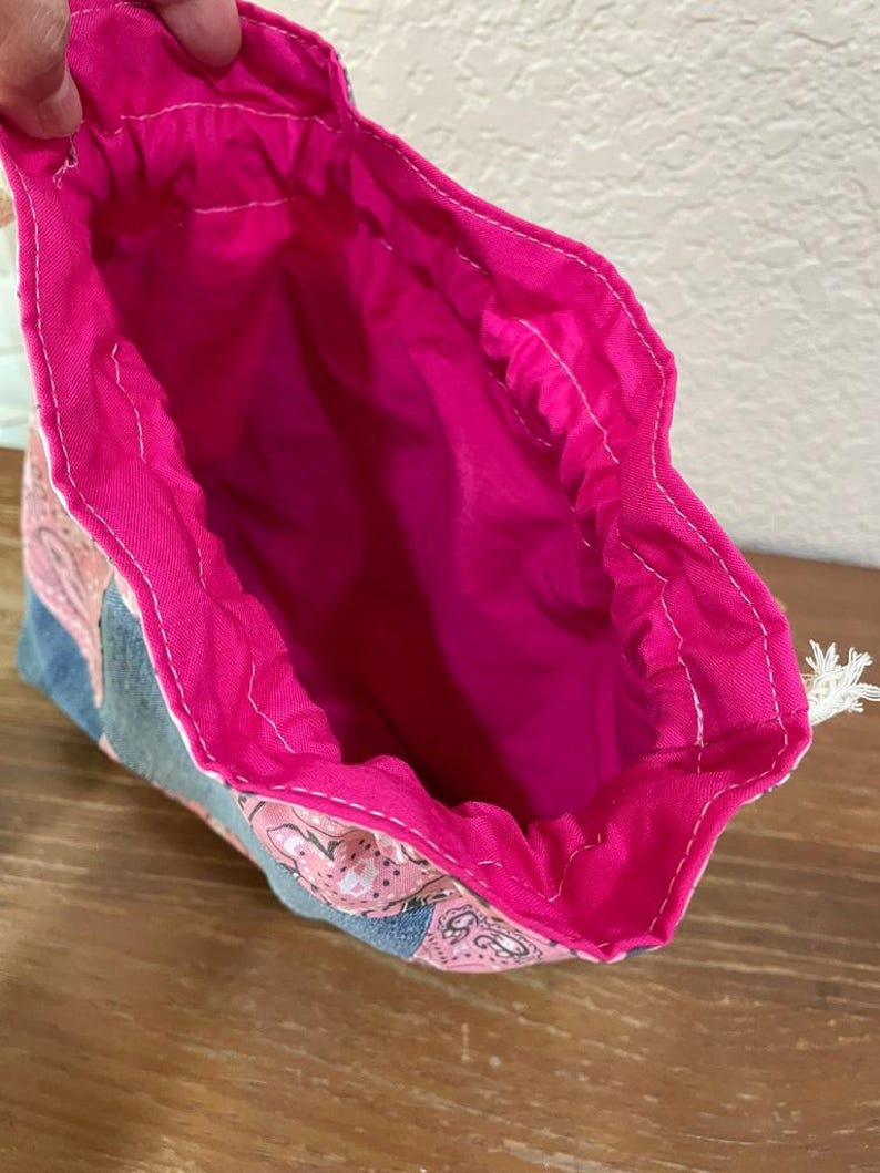 K&ouml;nnte beinhalten: Offener Kordelzugbeutel mit pinkfarbenem Innenraum und gemustertem Au&szlig;enbereich. Die Au&szlig;enseite weist ein paisleyartiges Design in Pink und Blau auf. Der Innenraum der Tasche ist leuchtend pink, und der Kordelzug ist wei&szlig;.