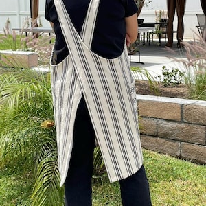May include: A cream-colored apron with vertical black stripes and two front pockets. The apron has a cross-back design and is worn over a black shirt and pants. The apron is made of a textured fabric.