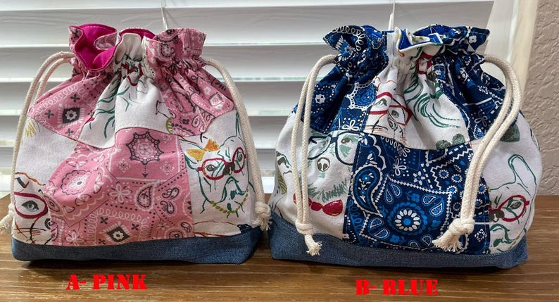 May include: Two drawstring bags with denim bottoms. One is pink with a paisley and cat print, the other is blue with a paisley and cat print. Both bags have a drawstring closure and a white cord. The text "A-PINK" and "B-BLUE" are visible.