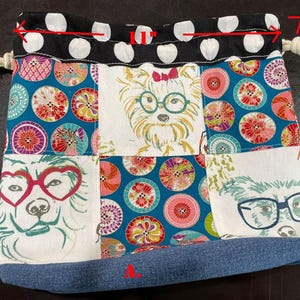 Pu&ograve; includere: Un sacco con coulisse patchwork, alto circa 23 cm e largo 2,5 cm, con illustrazioni di cani con gli occhiali. La borsa ha una base in denim, una parte superiore a pois bianchi e neri e una chiusura con coulisse.