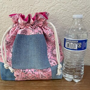 K&ouml;nnte beinhalten: Ein Kordelzugbeutel mit rosa Paisley-Muster und Jeans-Akzenten. Der Beutel hat einen wei&szlig;en Kordelzugverschluss und ein rosa Innenfutter. Eine transparente Plastikwasserflasche befindet sich neben dem Beutel.