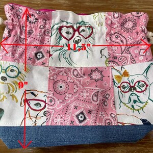 May include: A patchwork drawstring bag, approximately 11.5 inches wide and 9 inches tall, featuring dog illustrations wearing glasses and pink paisley patterns. The bag has a denim blue base and a pink interior.