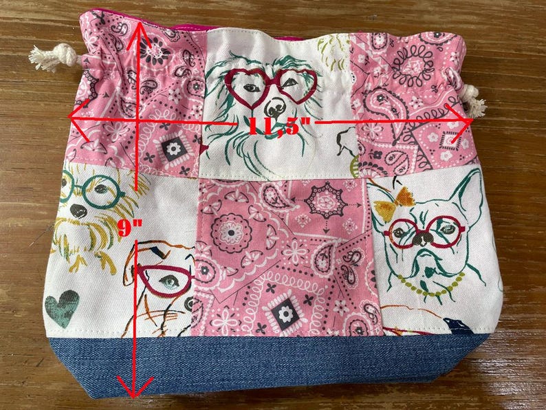 May include: A patchwork drawstring bag, approximately 11.5 inches wide and 9 inches tall, featuring dog illustrations wearing glasses and pink paisley patterns. The bag has a denim blue base and a pink interior.