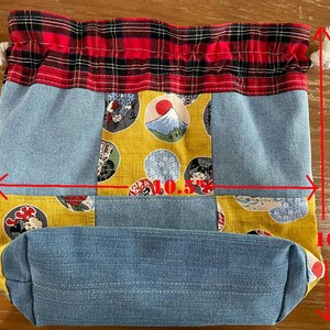 May include: A drawstring bag with a patchwork design. The bag is 10 inches tall and 10.5 inches wide. It features denim, yellow fabric with Japanese-style illustrations, and a red and black plaid top. White rope ties secure the top.