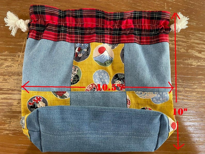 May include: A drawstring bag with a patchwork design. The bag is 10 inches tall and 10.5 inches wide. It features denim, yellow fabric with Japanese-style illustrations, and a red and black plaid top. White rope ties secure the top.