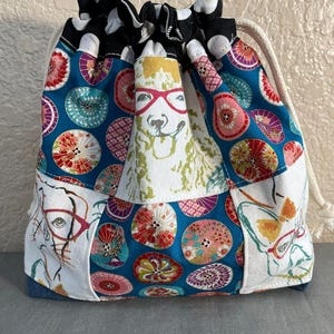 Pu&ograve; includere: Una borsa con coulisse con un design patchwork. La borsa presenta uno sfondo blu con motivi circolari colorati, pannelli bianchi con occhiali e un lama con gli occhiali. La parte superiore &egrave; nera con pois bianchi.