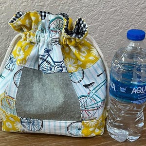 Op de afbeelding: Een trekkoordtas met een patchworkontwerp met fiets- en bloempatronen in geel, wit en blauw. De tas heeft een grijs denimachtig paneel en een zwart-wit geruite rand. Naast de tas staat een doorzichtige plastic waterfles met een blauwe dop.