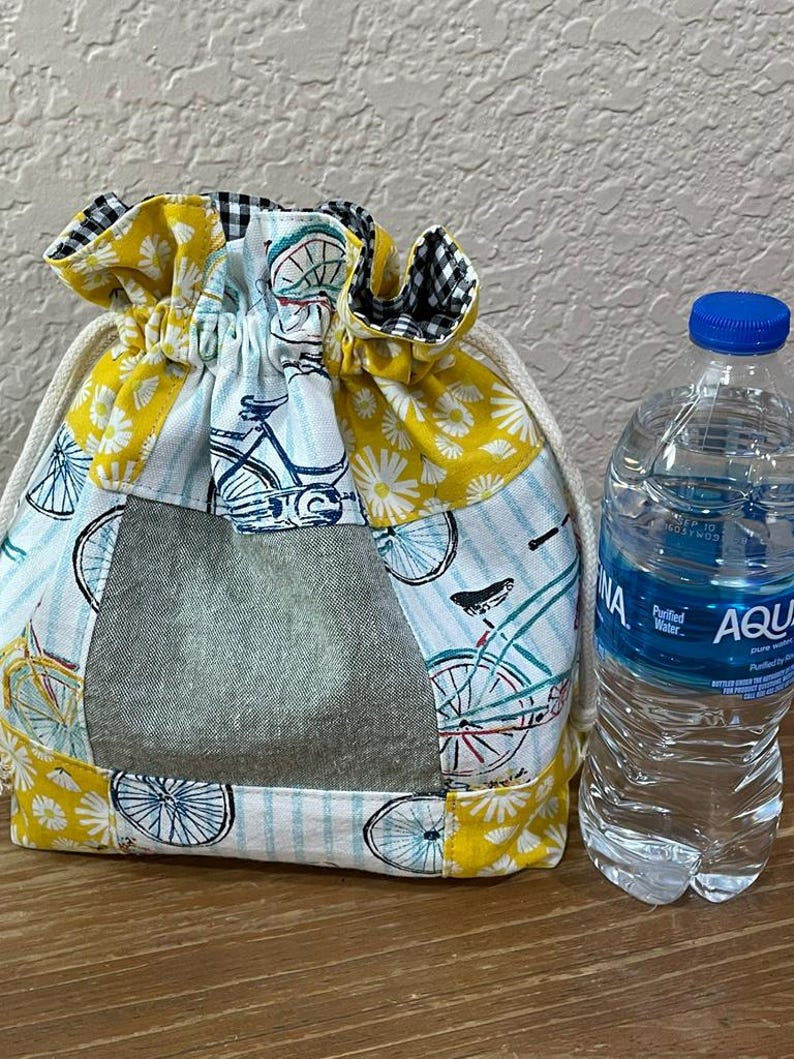 Op de afbeelding: Een trekkoordtas met een patchworkontwerp met fiets- en bloempatronen in geel, wit en blauw. De tas heeft een grijs denimachtig paneel en een zwart-wit geruite rand. Naast de tas staat een doorzichtige plastic waterfles met een blauwe dop.