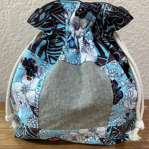 Op de afbeelding: Een trekkoordtas met een lichtblauw, bruin en wit bloemenpatroon, met vlinders en een grijze denim-achtige midden. De tas heeft een geplooid bovenstuk en witte trekkoorden.