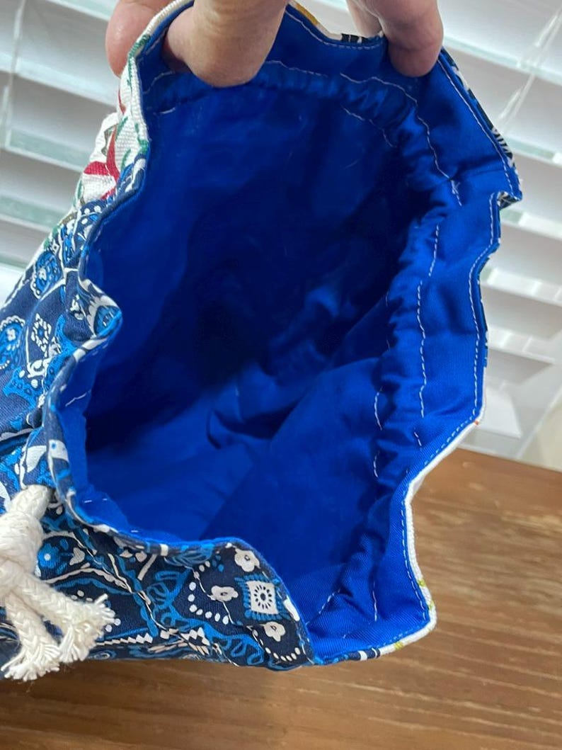May include: A blue drawstring bag with a white rope closure. The bag's exterior features a paisley pattern in shades of blue and white. The interior is a vibrant blue, and the bag is held open.