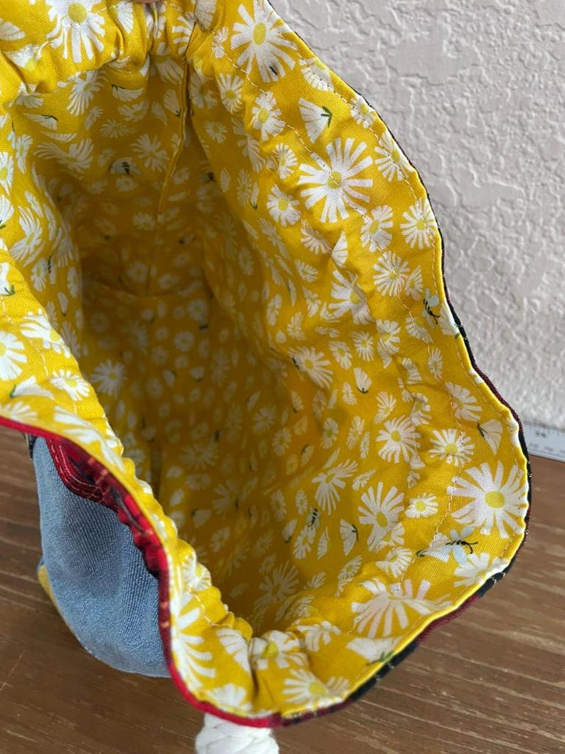 May include: A drawstring bag with a yellow daisy print lining. The exterior features denim, red plaid, and black fabric. A white drawstring is visible at the bottom. The bag's interior is bright yellow with white daisy flowers.