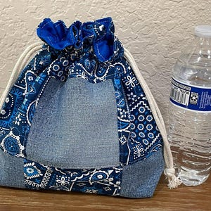 K&ouml;nnte beinhalten: Ein Kordelzugbeutel mit einem Denim- und blauen Paisley-Muster. Die Tasche hat einen Denim-Boden und einen blauen Stoffkordelzugverschluss. Eine transparente Plastikwasserflasche befindet sich neben der Tasche.