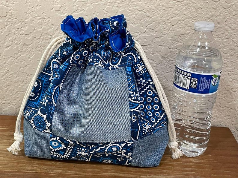 K&ouml;nnte beinhalten: Ein Kordelzugbeutel mit einem Denim- und blauen Paisley-Muster. Die Tasche hat einen Denim-Boden und einen blauen Stoffkordelzugverschluss. Eine transparente Plastikwasserflasche befindet sich neben der Tasche.