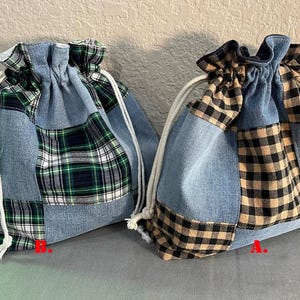 May include: Two denim drawstring bags with patchwork plaid accents. One bag features green, white, and navy plaid, while the other has black and tan plaid. Both bags have white drawstrings.