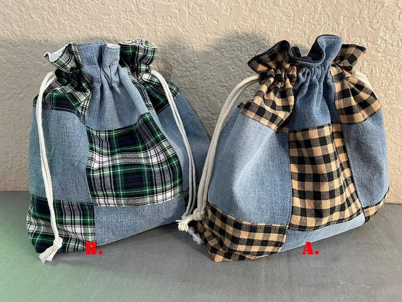 May include: Two denim drawstring bags with patchwork plaid accents. One bag features green, white, and navy plaid, while the other has black and tan plaid. Both bags have white drawstrings.