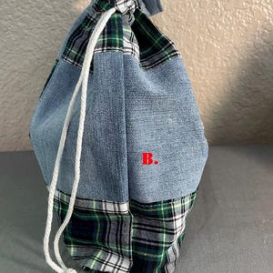 May include: A drawstring bag made from patchwork denim and plaid fabric. The bag features a light blue denim body with a green, black, and white plaid base and top. A white drawstring is used to close the bag.