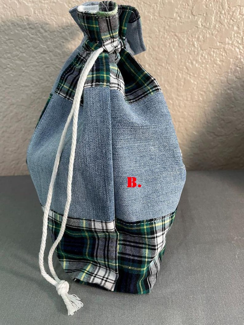 May include: A drawstring bag made from patchwork denim and plaid fabric. The bag features a light blue denim body with a green, black, and white plaid base and top. A white drawstring is used to close the bag.