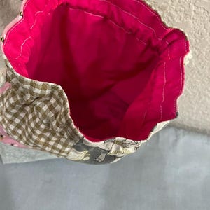 May include: Drawstring bag with a bright pink interior. The exterior has a patchwork design with brown and white gingham, floral, and grey patterned fabrics. Gold-coloured beads are sewn along the top edge.