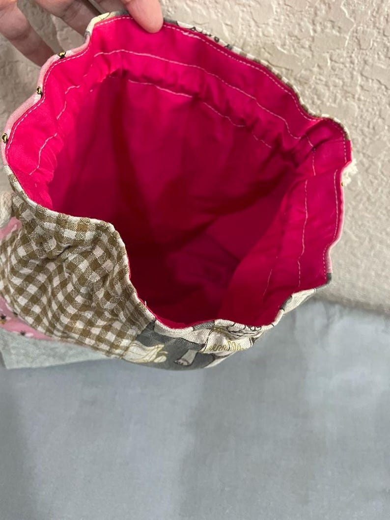 May include: Drawstring bag with a bright pink interior. The exterior has a patchwork design with brown and white gingham, floral, and grey patterned fabrics. Gold-coloured beads are sewn along the top edge.