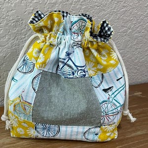 Op de afbeelding: Een trekkoordzakje met een patchwork-ontwerp. De tas heeft een denimachtig middenpaneel, omgeven door stof met fiets- en bloempatronen in geel, wit en blauw. De bovenrand is zwart-wit geruit en het trekkoord is wit.