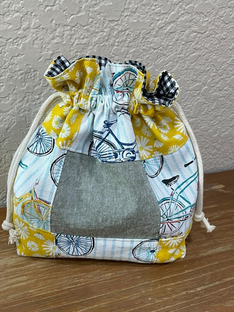 Op de afbeelding: Een trekkoordzakje met een patchwork-ontwerp. De tas heeft een denimachtig middenpaneel, omgeven door stof met fiets- en bloempatronen in geel, wit en blauw. De bovenrand is zwart-wit geruit en het trekkoord is wit.