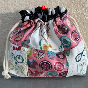 Pu&ograve; includere: Una borsa a coulisse patchwork con una variet&agrave; di motivi e colori. La borsa presenta un motivo floreale rosa, tessuto bianco con illustrazioni di cani e un bordo a pois bianchi e neri. Il cordoncino &egrave; una corda color crema.