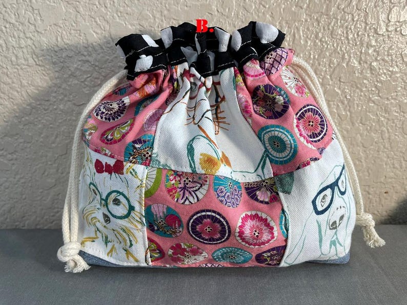Pu&ograve; includere: Una borsa a coulisse patchwork con una variet&agrave; di motivi e colori. La borsa presenta un motivo floreale rosa, tessuto bianco con illustrazioni di cani e un bordo a pois bianchi e neri. Il cordoncino &egrave; una corda color crema.