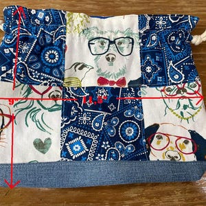 May include: A handmade drawstring bag, approximately 9 inches wide and 7 inches tall, featuring a patchwork design. The bag showcases dog illustrations wearing glasses, set against a blue paisley and white background. The base is denim.