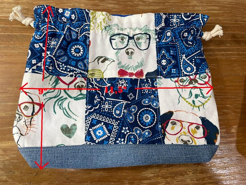 May include: A handmade drawstring bag, approximately 9 inches wide and 7 inches tall, featuring a patchwork design. The bag showcases dog illustrations wearing glasses, set against a blue paisley and white background. The base is denim.