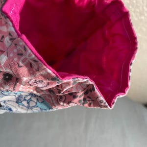 May include: A handmade fabric bag with a bright pink interior and a floral patterned exterior in shades of pink, white, and blue. The bag's interior is a solid, vibrant pink.