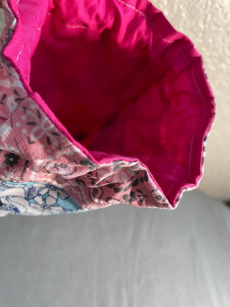 May include: A handmade fabric bag with a bright pink interior and a floral patterned exterior in shades of pink, white, and blue. The bag's interior is a solid, vibrant pink.