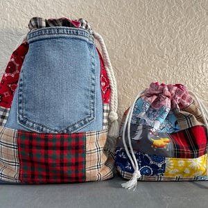 May include: Two drawstring bags. The larger bag is made from denim, red paisley, and plaid fabric. The smaller bag is made from a patchwork of colorful patterned fabrics. Both bags have white drawstring cords.
