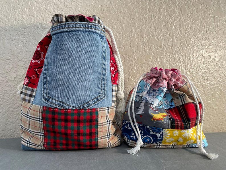 May include: Two drawstring bags. The larger bag is made from denim, red paisley, and plaid fabric. The smaller bag is made from a patchwork of colorful patterned fabrics. Both bags have white drawstring cords.