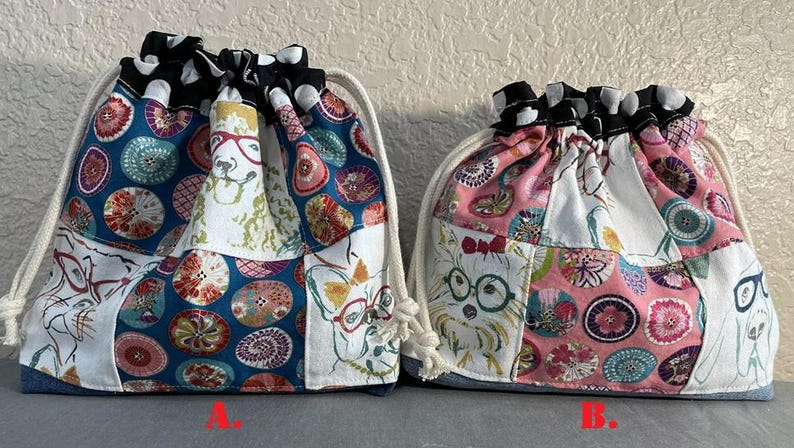 Pu&ograve; includere: Due sacchetti con coulisse con un design patchwork. Una borsa presenta uno sfondo blu con cerchi colorati e illustrazioni di cani con gli occhiali. L'altra borsa ha uno sfondo rosa con motivi simili e illustrazioni di cani. Entrambe le borse hanno un bordo a pois bianchi e neri.