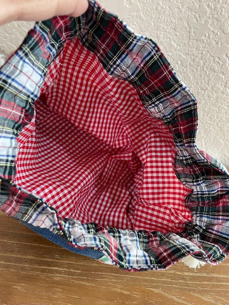 May include: A drawstring bag with a red and white gingham interior. The bag's exterior features a plaid pattern in red, green, black, and white. The bag is made of fabric and has a denim base.
